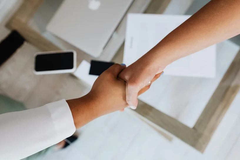 Overhead view of a professional handshaking with a client across a desk with blueprints and a smartphone.